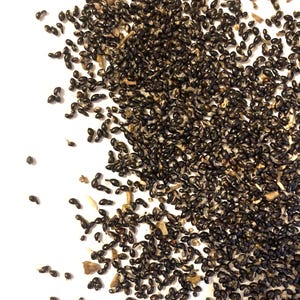 May include: A close-up shot of a pile of small, dark brown seeds scattered on a white surface. The seeds are oval-shaped and appear to be dry. Some small pieces of plant matter are also visible.