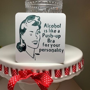 May include: White coaster with a green illustration of a woman winking and the text "Alcohol is like a Push-up Bra for your personality."