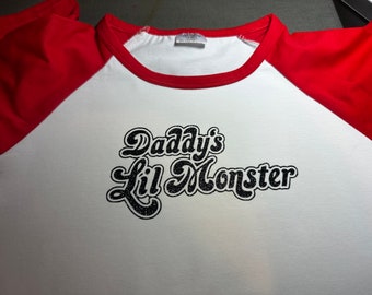 May include: A white baseball-style t-shirt with red raglan sleeves and a red crew neckline. The front of the shirt features the text "Daddy's Lil Monster" printed in a black, glittery, stylized cursive font across the center of the chest.