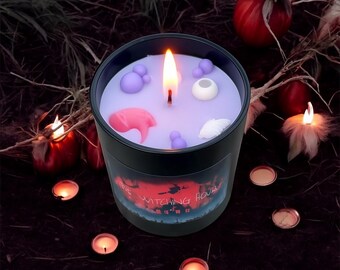 May include: A lit black candle with a purple wax surface, decorated with pink, white, and purple shapes. The candle has a label with the text "The Witching Hour" and is surrounded by small lit candles and decorative elements.