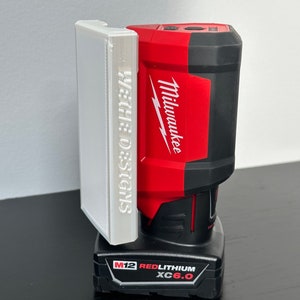 May include: A white 3D printed wall mount for a Milwaukee M12 RedLithium XC 6.0 battery. The mount has the text "Wattle Designs" printed on the side.