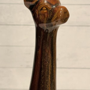 May include: A tall, brown ceramic giraffe figurine with a long neck and a small head. The giraffe has a glossy finish with shades of brown and tan. The figurine is set against a white background.