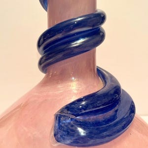 Puede incluir: Un jarr&oacute;n de vidrio rosa con un dise&ntilde;o de espiral de vidrio azul. El jarr&oacute;n tiene una base ancha y un cuello estrecho.