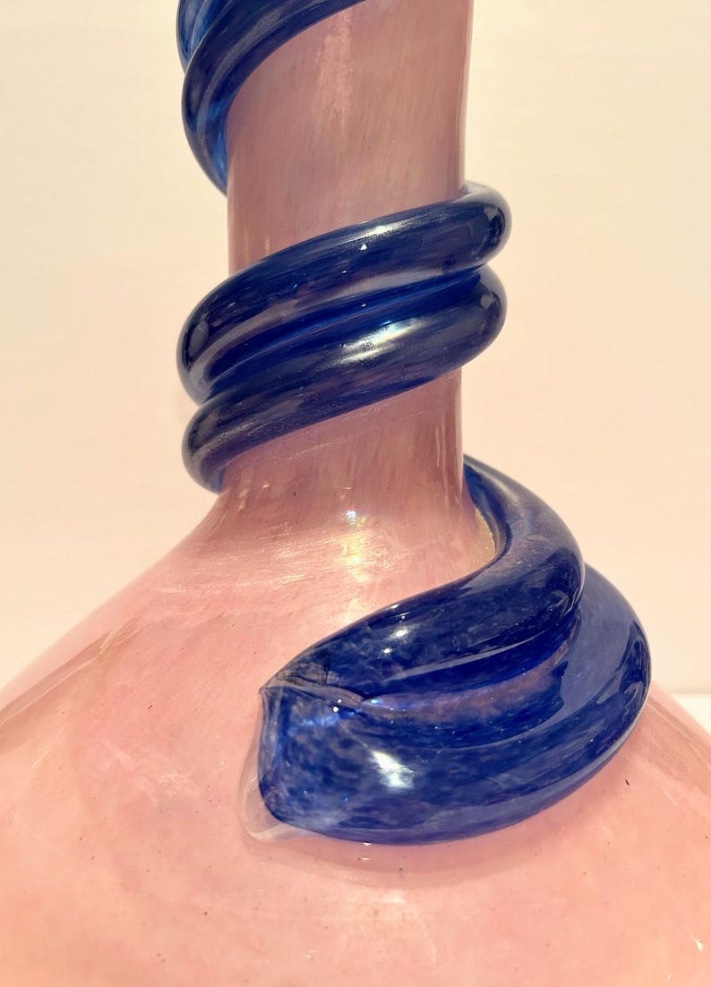 Puede incluir: Un jarr&oacute;n de vidrio rosa con un dise&ntilde;o de espiral de vidrio azul. El jarr&oacute;n tiene una base ancha y un cuello estrecho.