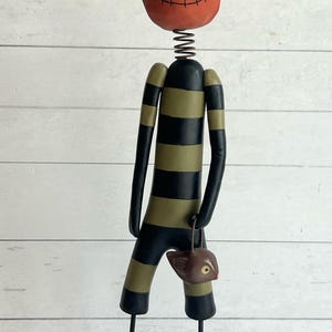 May include: A whimsical Halloween decoration featuring a pumpkin head with a black pointed hat. The body is striped black and olive green, with a spring neck and thin black legs. It holds a small brown bag. The figure stands on a brown rectangular base.