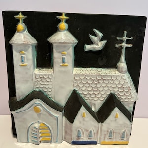 May include: A white ceramic wall plaque depicting a church with two towers, a dove flying above, and a cross on the roof. The church is painted with yellow, blue, and green accents against a black background.