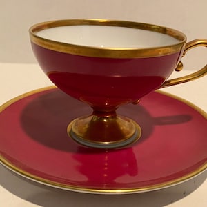 May include: A red and gold teacup and saucer set. The cup has a gold rim and a gold base. The saucer has a gold rim.