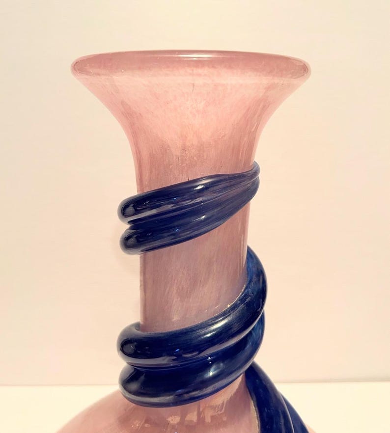 Puede incluir: Un jarr&oacute;n de vidrio rosa alto con una cinta de vidrio azul envuelta alrededor. El jarr&oacute;n tiene una base ancha y un cuello estrecho.