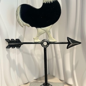 May include: A metal weather vane featuring a black and white rooster silhouette mounted on a horizontal arrow. The assembly is supported by a vertical black rod attached to a rectangular, speckled gray base with a distressed, weathered finish.