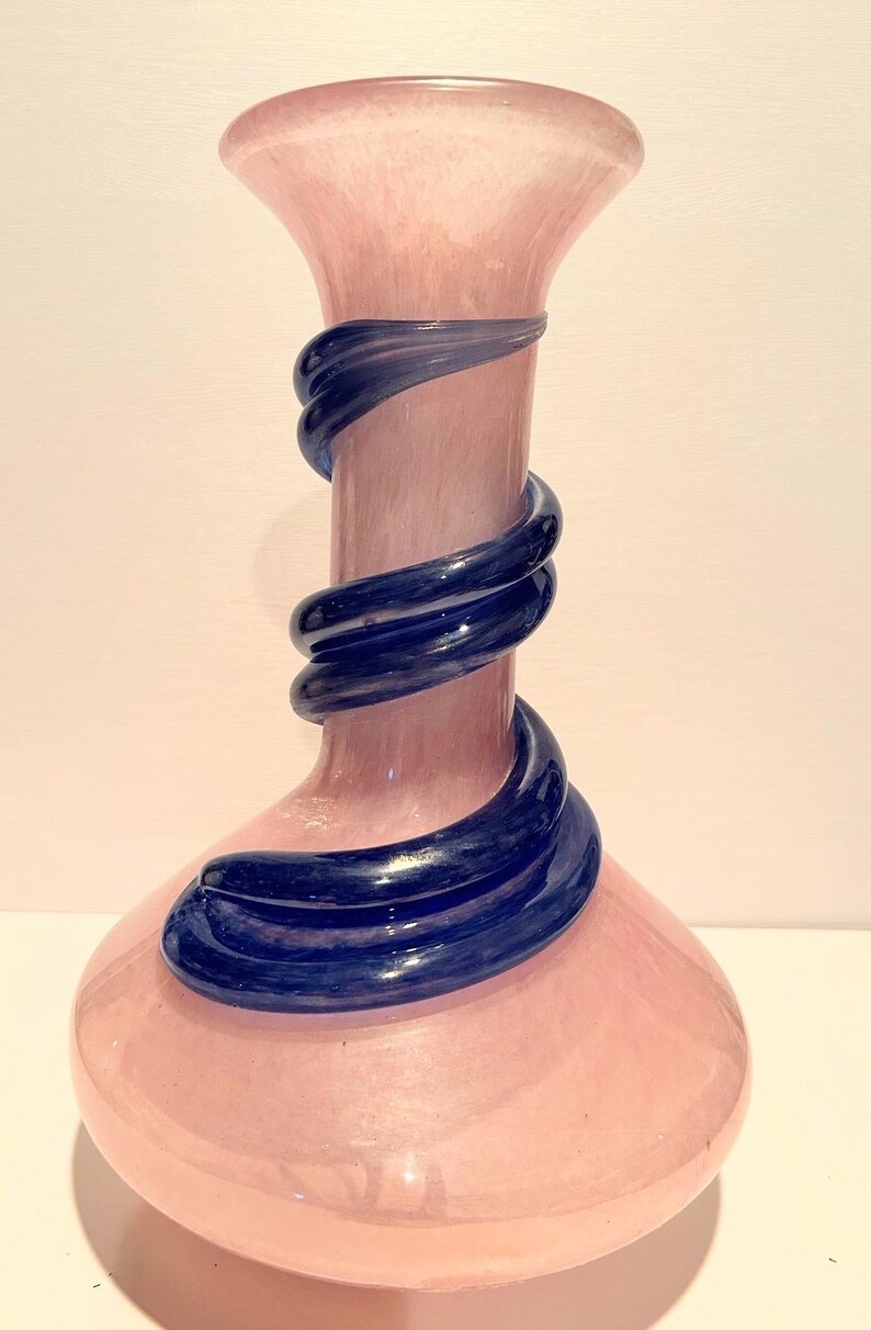 Puede incluir: Un jarr&oacute;n de vidrio rosa con un dise&ntilde;o de vidrio azul en espiral. El jarr&oacute;n tiene una base ancha y un cuello estrecho.