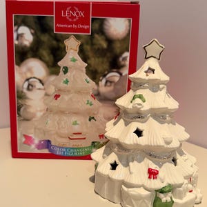 May include: A white ceramic Christmas tree figurine featuring a gold-trimmed star topper, silver glitter accents, and molded decorations including red bows and green gifts. The packaging box displays the Lenox brand and the text Color Changing Lit Figurine.