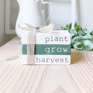 May include: Three wooden blocks stacked on top of each other. The blocks are painted white and green. The top block says "plant" in white lettering. The middle block says "grow" in white lettering. The bottom block says "harvest" in white lettering. The blocks are tied together with a brown ribbon.