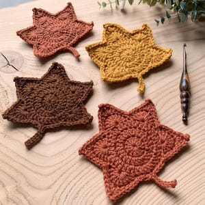 May include: Four handmade crochet leaf coasters in shades of brown and yellow, arranged on a wooden surface. A crochet hook with a wooden handle rests nearby. The coasters are in various autumnal colors.