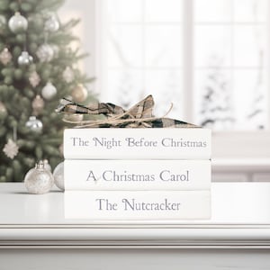 May include: Three white books stacked, with titles "The Night Before Christmas", "A Christmas Carol", and "The Nutcracker". A plaid ribbon and twine are tied on top. A Christmas tree and ornaments are in the background.