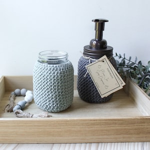May include: Two crocheted mason jar covers, one in grey and one in green, on a wooden tray. The grey jar has a brown pump top and a tag that reads "Handmade Mason Jar Soap Dispenser".
