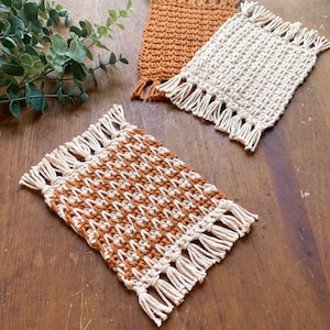 May include: Three handmade macrame coasters in shades of tan and brown. One coaster features a geometric pattern, while the others are solid colors. Each coaster has a fringe detail along the edges, adding a decorative touch.