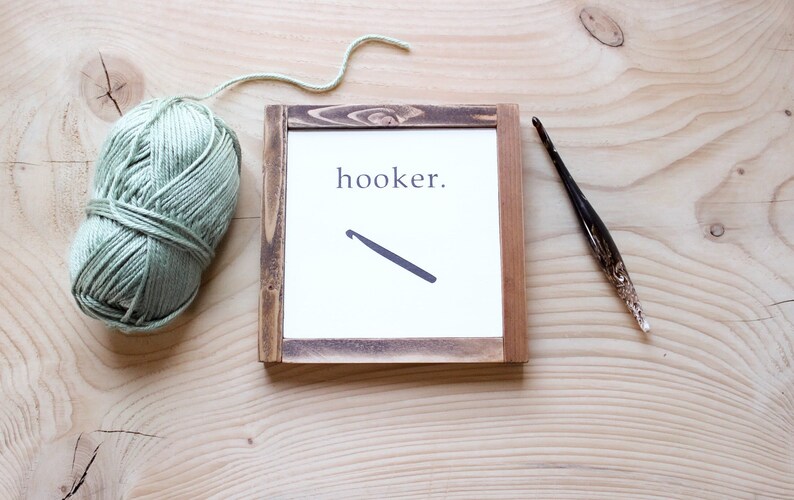 Może przedstawiać: Ramka z napisem "hooker." i ilustracją szydełka. Kulka zielonej wł&oacute;czki i szydełko leżą na drewnianej powierzchni.