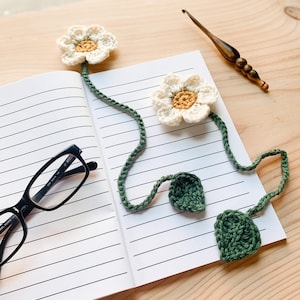 May include: A crocheted daisy bookmark with cream petals and a yellow center rests on a lined notebook. The green stem and leaves extend from the flower. Black eyeglasses are on the left side of the notebook. A wooden crochet hook is on the right.