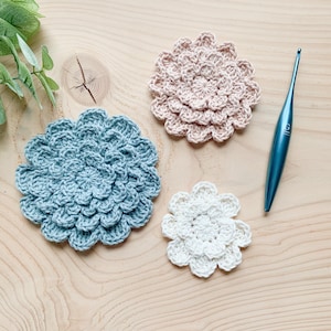 May include: Three crocheted flower-shaped coasters in blue, pink, and white, with a blue crochet hook. The coasters are displayed on a light wooden surface, highlighting a handmade craft theme.