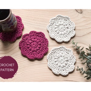 May include: Three crocheted coasters, two in a light beige color and one in a deep burgundy color. The coasters are on a wooden surface with a sprig of greenery in the background. The text "CROCHET PATTERN" is written in white on a burgundy circle.