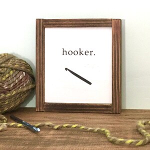 Może przedstawiać: Drewniany znak z napisem "hooker" i grafiką haczyka do rob&oacute;tek ręcznych. Znak stoi na drewnianej powierzchni z wł&oacute;czką i haczykiem do rob&oacute;tek ręcznych.