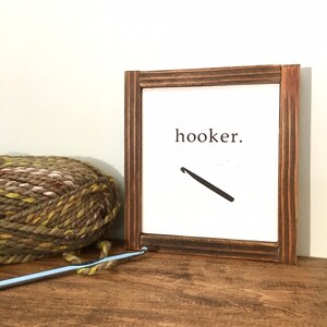 Może przedstawiać: Drewniany znak w ramie z napisem "hooker" i grafiką haczyka do rob&oacute;tek ręcznych. Znak stoi na drewnianej powierzchni z kłębkiem wł&oacute;czki i haczykiem do rob&oacute;tek ręcznych.