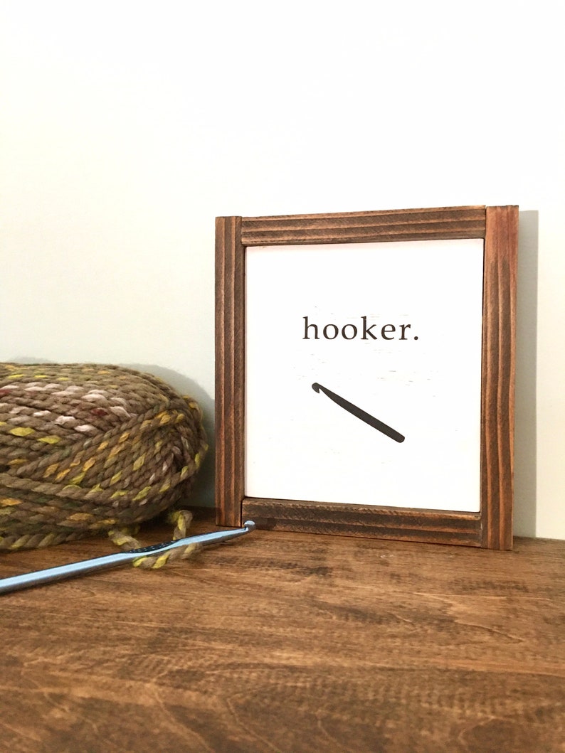 Może przedstawiać: Drewniany znak w ramie z napisem "hooker" i grafiką haczyka do rob&oacute;tek ręcznych. Znak stoi na drewnianej powierzchni z kłębkiem wł&oacute;czki i haczykiem do rob&oacute;tek ręcznych.