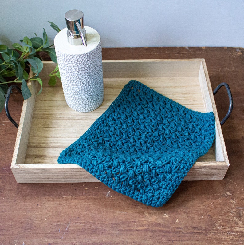 May include: A wooden tray with black metal handles holds a teal crocheted dishcloth and a white ceramic soap dispenser with a silver pump.