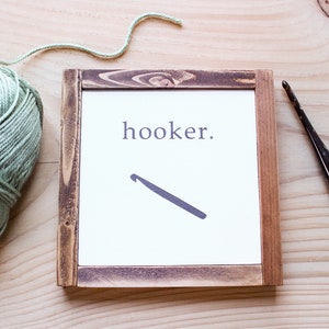 Może przedstawiać: Ramka z napisem "hooker." i sylwetką szydełka. Kulka zielonej wł&oacute;czki i szydełko leżą na drewnianej powierzchni.