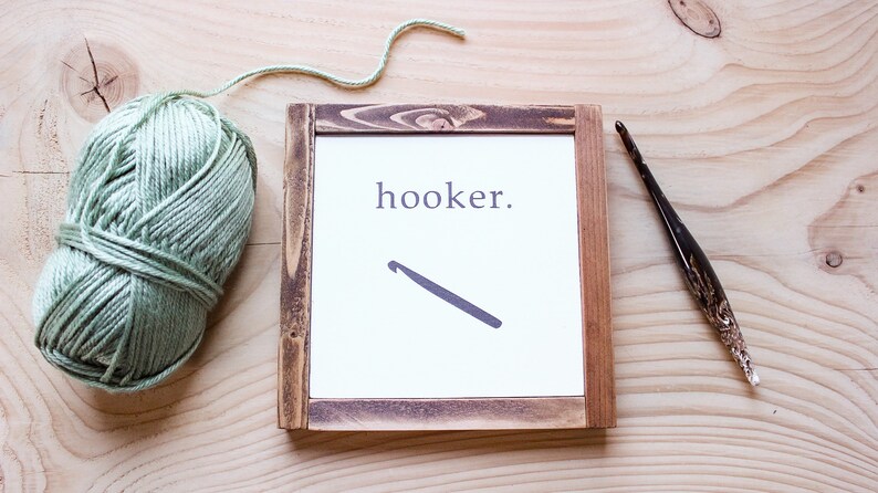 Może przedstawiać: Ramka z napisem "hooker." i sylwetką szydełka. Kulka zielonej wł&oacute;czki i szydełko leżą na drewnianej powierzchni.