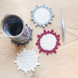 May include: Handmade crochet coasters in white, light blue, and burgundy. The coasters have a sunburst design and are placed on a wooden surface. A ceramic mug and a silver tool are also present.