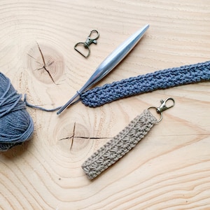 May include: A close-up of a crocheted keychain with a silver hook and a ball of gray yarn. The keychain is made with a beige yarn and has a silver clasp.