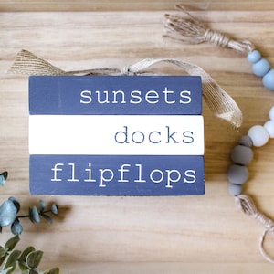 May include: Three wooden blocks stacked on top of each other. The top block is dark blue with the word "sunsets" in white lettering. The middle block is white with the word "docks" in black lettering. The bottom block is dark blue with the word "flipflops" in white lettering. A strand of gray, white, and blue beads with a brown tassel hangs to the right of the blocks.