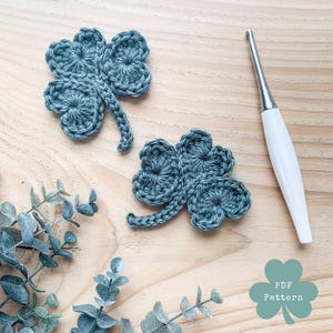 May include: Two crocheted shamrocks in a muted teal color, with detailed leaf patterns and stems, are displayed on a light wood surface. A crochet hook with a white handle and silver metal tip is positioned nearby. The image also includes sprigs of greenery.