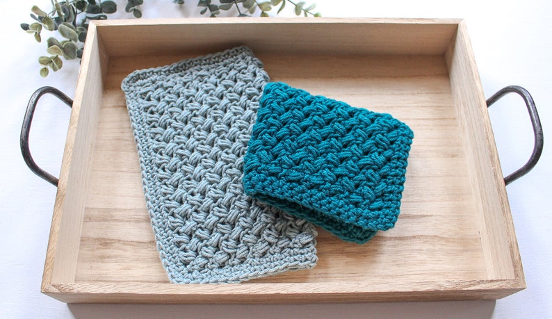 May include: Two crocheted dishcloths in a wooden tray. One dishcloth is teal blue and the other is light grey.