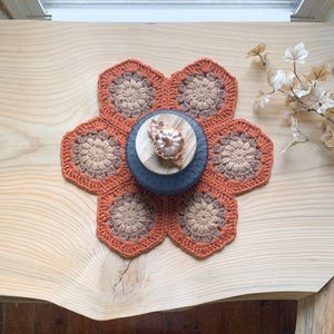 May include: A handmade, orange and tan crochet flower-shaped placemat. A dark blue, wooden bowl with a small, copper-colored decorative object sits in the center. The placemat is on a light-colored wooden surface.