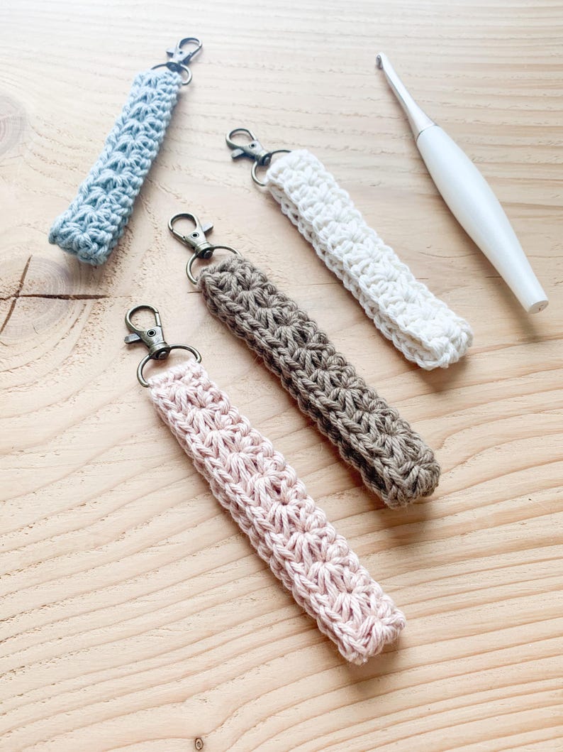 May include: Four crocheted keychains in different colors: light blue, white, brown, and pink. Each keychain has a metal clasp and is made with a delicate stitch pattern.
