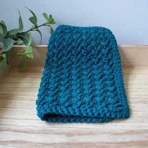 May include: A teal blue knitted dishcloth with a textured pattern. The dishcloth is sitting on a wooden surface.