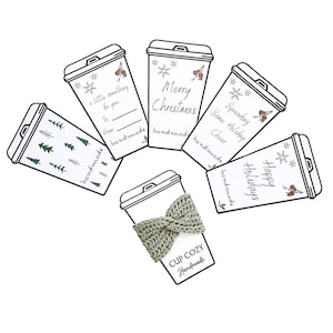 May include: A collection of white paper cup-shaped gift tags with black outlines. The tags feature holiday-themed text like "Merry Christmas" and "Happy Holidays", along with snowflake and pinecone illustrations. One tag has a knitted cup cozy.