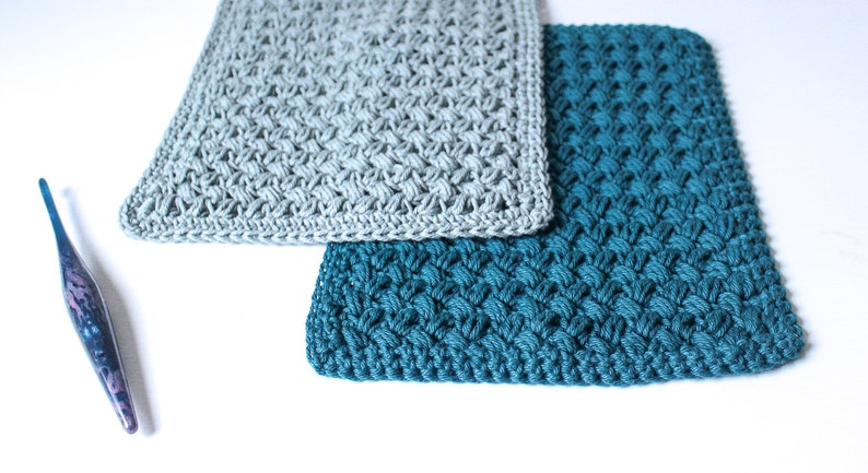 May include: Two crocheted squares, one in a light grey colour and one in a teal blue colour. The squares are made with a textured stitch pattern.