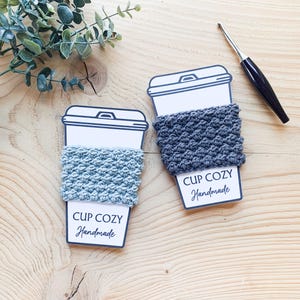 May include: Two paper coffee cup cutouts with crocheted cozies in light blue and grey. Each cup has a label that reads "CUP COZY Handmade". A crochet hook rests on the wooden surface, along with a sprig of greenery.