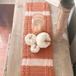 以下が含まれることがあります： オレンジと白の手作りのかぎ針編みのテーブルランナー、キャンドルと3つの白いニットカボチャ。木製の看板には「lets stay home」と書かれています。アイテムは、明るい茶色の木製のテーブルに配置されています。