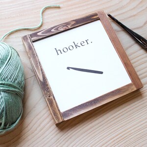 Może przedstawiać: Ramka z napisem "hooker." i czarną sylwetką szydełka. Kulka zielonej wł&oacute;czki i szydełko leżą na drewnianej powierzchni.