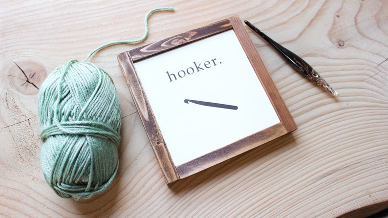 Może przedstawiać: Ramka z napisem "hooker." i czarną sylwetką szydełka. Kulka zielonej wł&oacute;czki i szydełko leżą na drewnianej powierzchni.