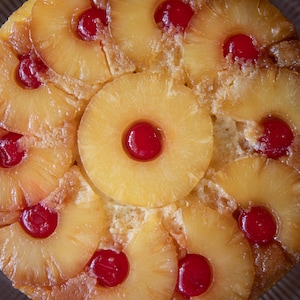 Può includere: Una torta rovesciata all'ananas con una crosta dorata, guarnita con anelli di ananas e ciliegie al maraschino. La torta è presentata su un'alzatina di vetro trasparente, un dessert classico.