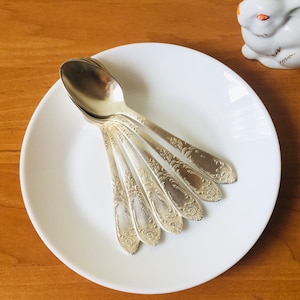 May include: A set of seven silver teaspoons with ornate designs on a white plate. The spoons are arranged in a fan shape.