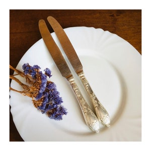 Pode incluir: Duas facas de prata com cabos ornamentados são colocadas em um prato branco com um pequeno buquê de flores secas roxas.