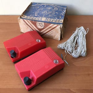 以下が含まれることがあります： 黒いノブと透明なライトが付いた、ヴィンテージの赤いモールス信号練習セット。キリル文字と図が書かれた箱がセットの後ろにあり、白いワイヤーの束も付いています。箱は青と茶色です。