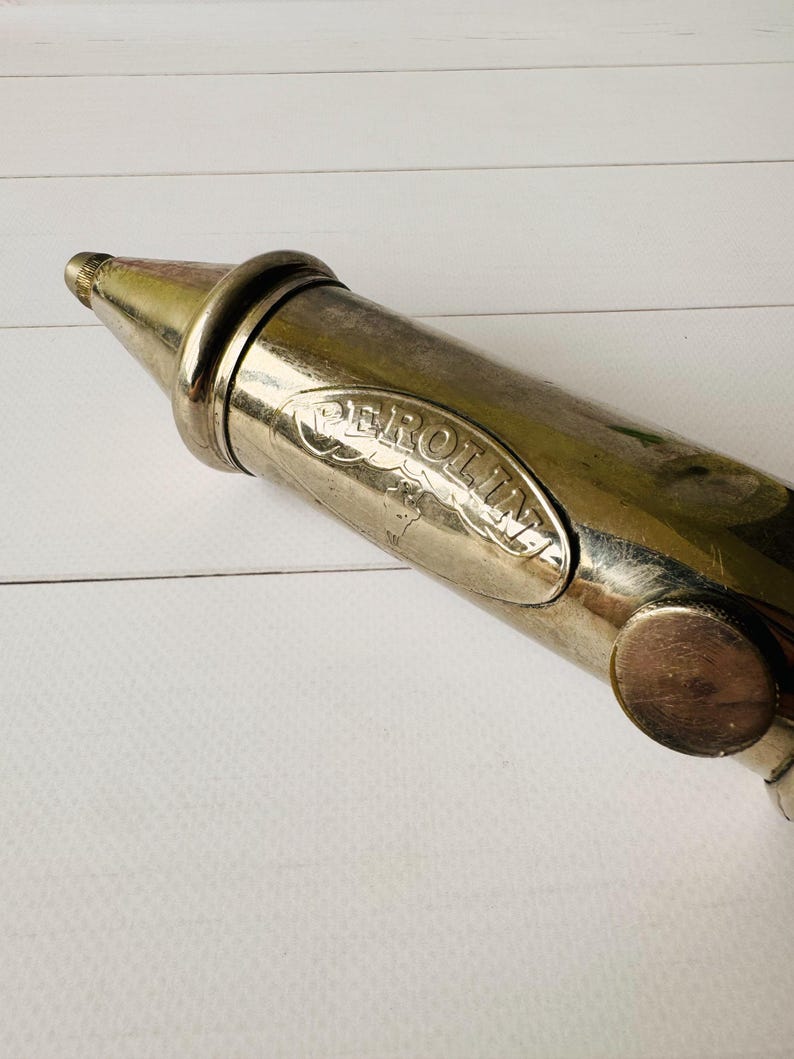 Close-up of the Perolin brand mark on a vintage air pump, showing the embossed logo on the metal body of an antique grease pump with aged surface