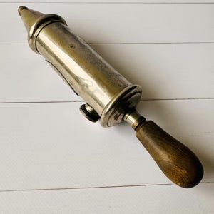 Vintage Perolin grease gun shown at an angle, antique tool with worn metal surface and wooden handle, ideal for industrial decor or man cave decor
